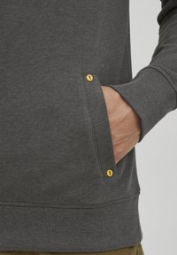 Sudadera gris con un acabado texturizado, que presenta un bolsillo lateral con detalles en hardware amarillo y puños y dobladillo acanalados.