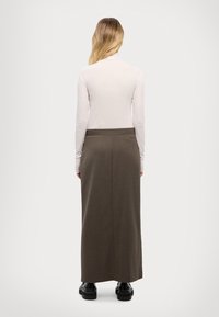 Donna con lunghi capelli biondi, che indossa una maglietta bianca a maniche lunghe, una gonna marrone lunga fino alla caviglia e scarpe nere spesse, in piedi con le spalle rivolte verso di noi.