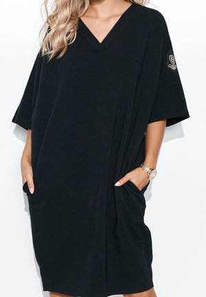 Robe T-shirt noire surdimensionnée avec col en V, manches courtes, poches latérales et un emblème décoratif sur la manche gauche. Tissu doux et léger.