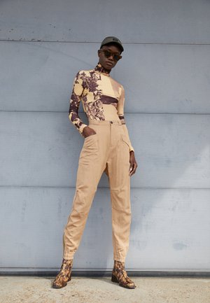 Bodysuit de manga longa em padrão abstrato amarelo e castanho, calças de cintura alta beje e botas de tornozelo castanhas com padrão.