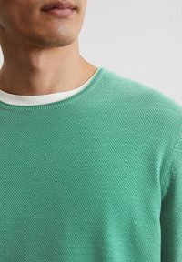 Hellgrüner, strukturierter Pullover mit Rundhalsausschnitt, der eine weiße Innenlage am Kragen hat, eng anliegend geschnitten ist und aus weichem Material besteht.
