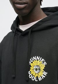 Felpa nera con cappuccio con una grafica circolare che raffigura un girasole sorridente con occhiali da sole, testo "Sunnie's Pool Bar" in lettere bianche in grassetto.