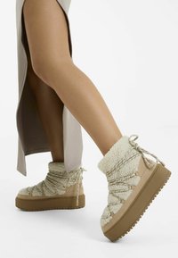 Beige plateau-støvler med blødt shearling-overmateriale, teksturerede snørebånd og metal-accenter. Har en klodset gummisål for godt greb.