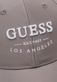 Hallikas müts valge tikitud "GUESS" logo, "EST.1981" ja "LOS ANGELES." Siledat kangast, struktureeritud kuju ja tikandid on hästi nähtavad.