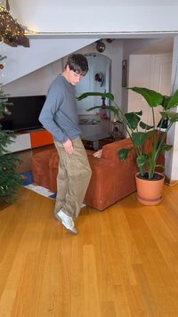 Jeune homme en vêtements décontractés, en équilibre sur un pied dans un salon avec un sol en bois, un canapé orange, une grande plante en pot et un sapin de Noël décoré.