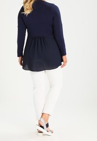 Haut bleu marine à manches longues avec un dos plissé, associé à un pantalon blanc longueur cheville et des sandales blanches avec des accents bleu marine.