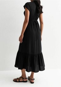 Svart maxiklänning i mjukt tyg, med knytbart midjeband, korta ärmar och en lager-på-lager volangnedre del. Matchad med svarta flata sandaler.