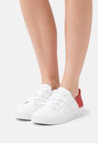 Baskets blanches avec accents rouges, design à lacets, semelle en caoutchouc blanc texturé, détails perforés sur les côtés et dessus en synthétique lisse.
