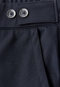 Tmavý pás látky s dvěma kovovými knoflíky vyrytými nápisem „Street One“, s detailním šitím a plisovanou částí pod knoflíky.