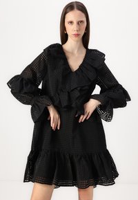 Svart rufsete kjole med V-hals, teksturerte mønstre, lange klokkeermer og en flagrende kant. Laget av lett stoff med gjennomsiktige detaljer.