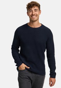Maglione a maniche lunghe in a nido d'ape di colore blu navy con scollo a coste e finitura testurizzata, abbinato a pantaloni grigi scuri a gamba affusolata, con le mani nei taschini.