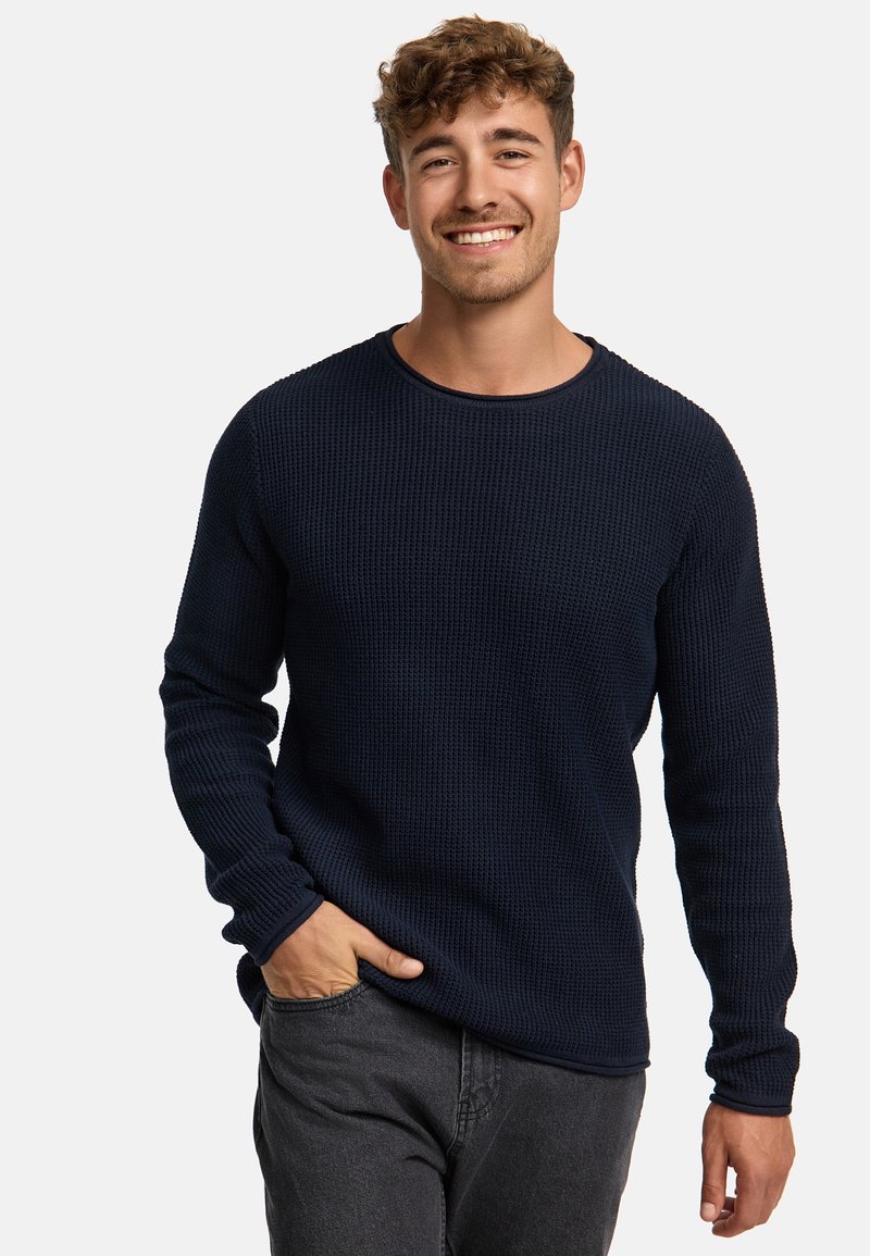 Maglione a maniche lunghe in a nido d'ape di colore blu navy con scollo a coste e finitura testurizzata, abbinato a pantaloni grigi scuri a gamba affusolata, con le mani nei taschini.