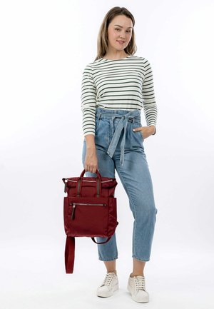 Roter Stoff-Rucksack mit zwei oberen Griffen, verstellbaren Trägern und mehreren Reißverschlusstaschen, kombiniert mit blauen hochtaillierten Jeans.