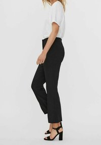 Pantalones negros ajustados con corte recto, combinados con una blusa blanca suelta. La modelo lleva sandalias negras de tacón bloque. Diseño simple y moderno.