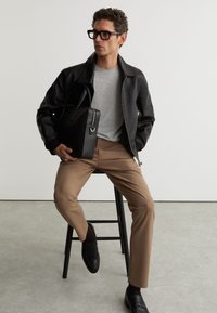 Homme portant une veste en cuir noire, une chemise grise, un pantalon beige, des chaussures noires et des lunettes, assis sur un tabouret tenant un sac en cuir noir.