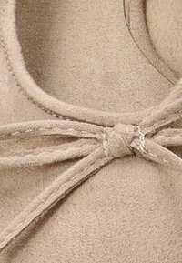 Zapato de ante beige con punta redonda y bordes cosidos. Los detalles incluyen un lazo en la parte delantera, que resalta la textura del material.