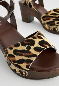Sandale cu platformă cu imprimeu leopard, având o bază din piele maro, cu o curea mai lată și cu catarame decorative din aur pe margine.