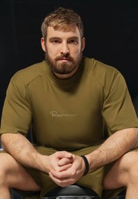 Chemise militaire verte à manches courtes avec un col rond, présentant un logo blanc, assortie à un short. Texture lisse et coupe décontractée.