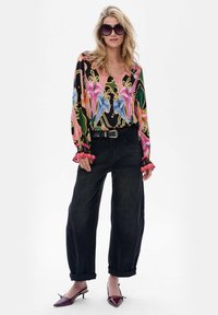 Kobieta w dużych okrągłych okularach przeciwsłonecznych, kwiecistej bluzce z długim rękawem, czarnych szerokich jeansach z paskiem oraz bordowych szpilkach w szpic.