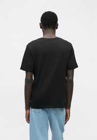 Uomo con capelli corti e ricci che indossa una t-shirt nera semplice e jeans azzurri chiari, di spalle su uno sfondo bianco.