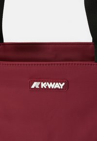 Bordeaux tas met een gladde textuur, met een witte "K-WAY" logo-label en zwarte banden. Eenvoudig ontwerp, geen extra patronen of accenten.