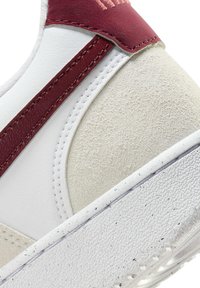 White sneaker with a textured white rubber sole, featuring red leather accents and a soft beige suede panel. Visible stitching details.