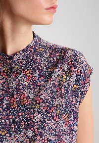 Blusa floral con fondo azul marino, que presenta pequeñas flores rosas, amarillas y blancas. Mangas cortas tipo capucha y escote redondo. Detalle de botones presente.