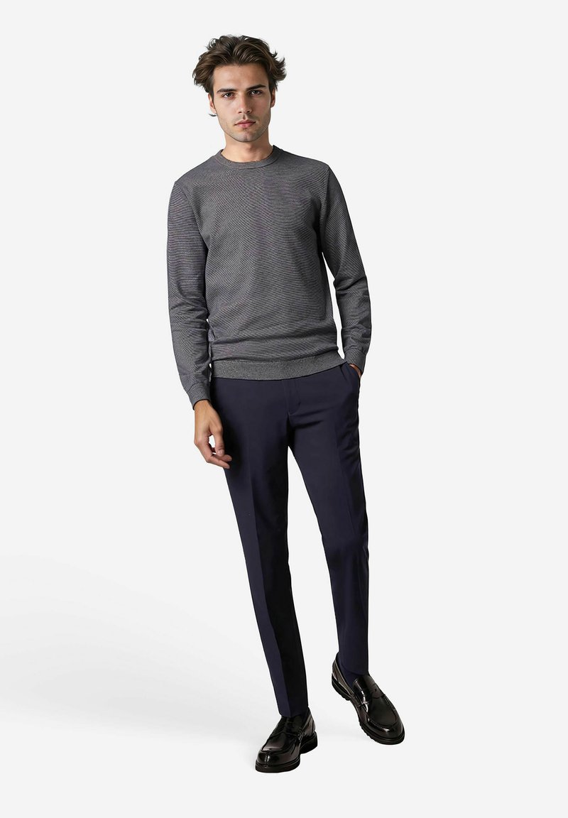 Grauer Strickpullover mit einem strukturierten Muster, kombiniert mit dunkelblauen Hosen und schwarzen Schuhen, stehend vor einem weißen Hintergrund.