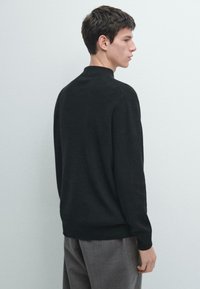 Schwarzer Strickpullover mit hohem Kragen, gerippten Bündchen und Saum. Glatte Textur, die eine klassische Silhouette mit zurückhaltenden Designdetails zeigt.