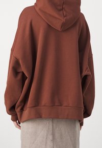Osoba v oversize hnědém svršku s kapucí a světlé šedé sukni, zobrazená zezadu na jednobarevném pozadí.