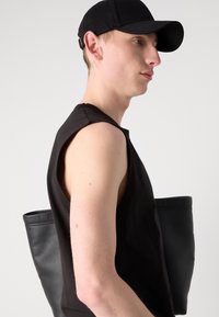 Tricou negru fără mâneci și șapcă neagră purtate de o persoană care poartă o geantă de umăr din piele neagră. Texturi netede și design simplu.