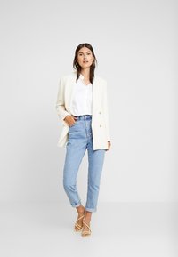 Weiße doppelt knöpfbare Blazer, über einem weißen Hemd getragen, kombiniert mit hellblauen hochgeschnittenen Jeans und gelben Riemchenheel.
