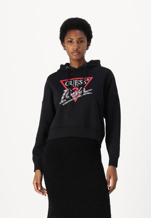 Black cropped hoodie with a red triangle logo. Features a drawstring hood and a soft texture. Worn over a black fuzzy skirt.