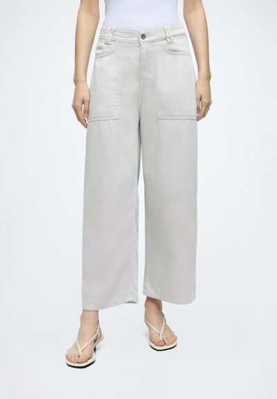 Personne portant un pantalon large gris clair avec poches plaquées à l'avant, un haut blanc sans manches et des sandales blanches type tongs, debout devant un fond uni.