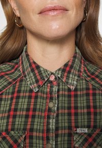 Camicia a quadri verde, nera e rossa con colletto, dotata di un bottone rosa e di un'etichetta con il logo sulla tasca laterale.