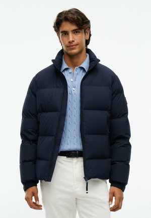 Joven con chaqueta acolchada azul marino sobre un suéter azul claro de punto trenzado y pantalones blancos, de pie frente a un fondo liso.