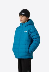 Tengeri zöld színű steppelt dzseki kapucnival, elöl cipzárral és oldalzsebekkel. Tartozik hozzá egy fekete kötött sapka logóval.