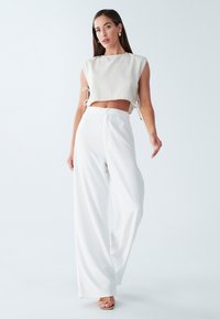 Beskåret, lys beige top med sidelukninger; hvide, vide bukser med vertikal tekstur; model iført nude hæle, minimale accessories.