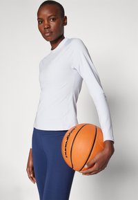 Hvit langermet idrettsgenser og marineblå leggings, holder en oransje basketball, viser glatt stofftekstur og atletisk passform.