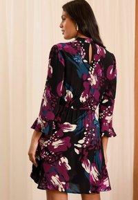 Rochie neagră cu modele florale colorate în nuanțe de mov, albastru și alb. Prezintă mâneci evazate, un decolteu cu orificiu în spate și o talie strânsă.