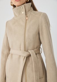 Beige fleece takki korkealla kauluksella, kultaisella vetoketjulla ja yhteensopivalla vyöllä. Huomionarvoisia yksityiskohtia ovat napit ja sivutaskut. Pehmeä rakenne.