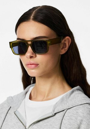 Mujer con cabello largo y oscuro que lleva gafas de sol rectangulares grandes de color verde oliva y una sudadera con capucha gris claro con cremallera sobre una camiseta blanca.