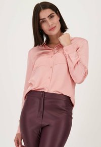 Lichtroze, knoopsluiting blouse met lange mouwen en borstzakken, gecombineerd met strakke, donkerburgundy, glanzende leren broek.