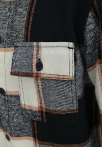 Chemise en flanelle à motif à carreaux avec des accents gris, blanc et orange. Elle possède une poche poitrine avec des boutons foncés et une texture douce.