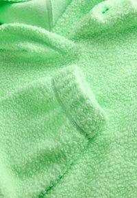 Hanorac din fleece de culoare verde deschis, cu o suprafață texturată și pufoasă. Dispune de glugă și manșete strânse, cu detalii de cusut vizibile pentru durabilitate.