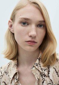 Close-up van een persoon met lichtblond, schouderlang haar, die een beige shirt met een zwart slangpatroon draagt. Neutrale achtergrond.