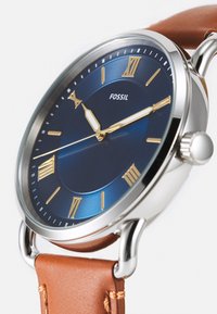 Montre Fossil avec cadran analogique bleu, chiffres romains dorés, boîtier en acier inoxydable argenté et bracelet en cuir brun.