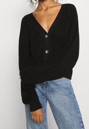Kvinde iført en sort ribstrikket cardigan med store knapper og løstsiddende ærmer, kombineret med lyseblå denimjeans.