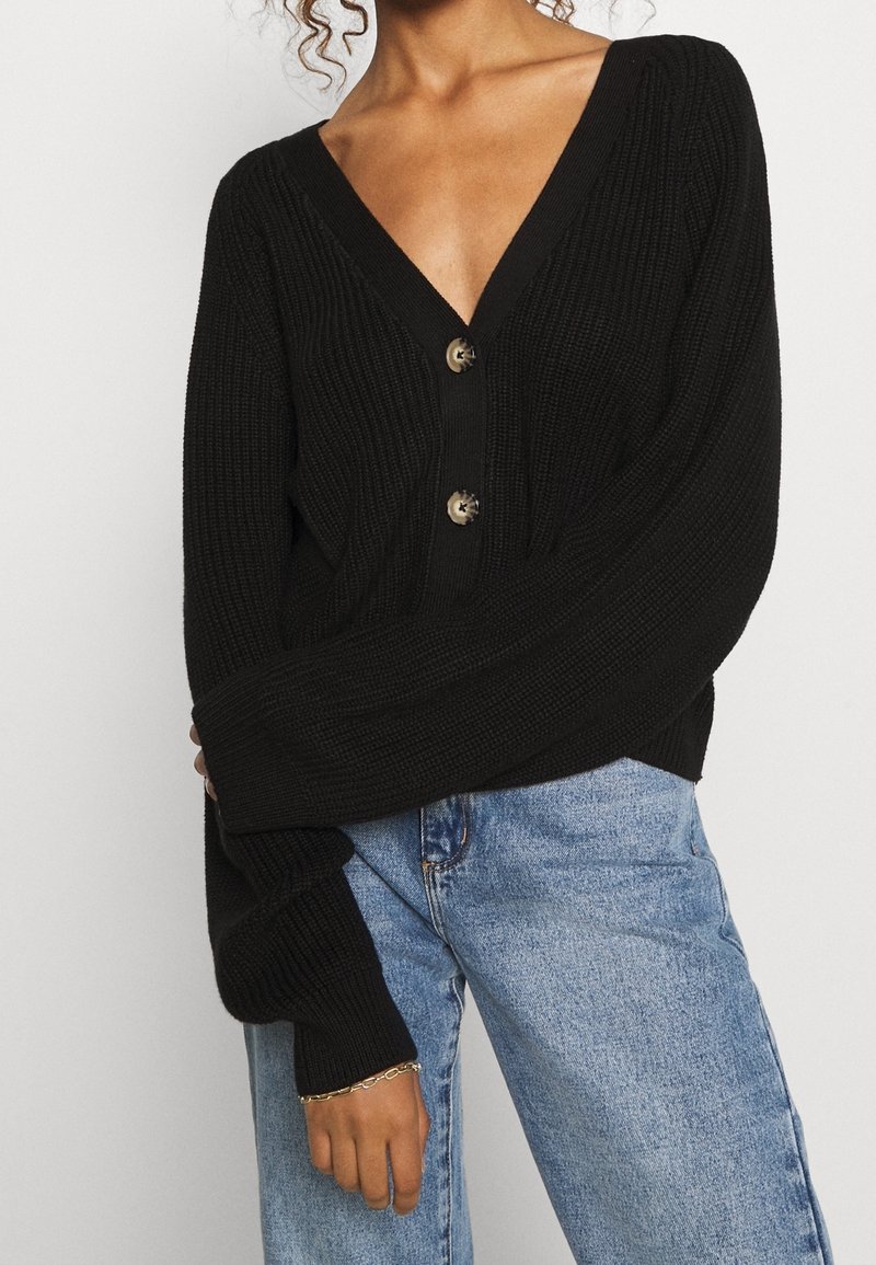 Femme portant un cardigan noir en tricot côtelé avec de gros boutons et des manches larges, associé à un jean en denim bleu clair.