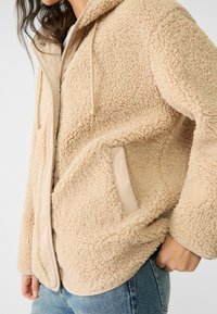 Beżowa sztuczna kurtka z futerkiem, z teksturowaną powierzchnią zewnętrzną, kapturem i kremowymi akcentami. Posiada boczne kieszenie oraz zamek błyskawiczny.